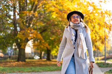 Beautiful middle-aged woman wearing jeans, gray coat, checkered warm scarf, black brimmed hat and sunglasses walking along sidewalk surrounded by big trees in city on sunny autumn day. Front view