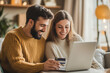 © Benjawan - Happy couple with credit card in home using laptop for shopping.