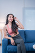 © Tj - Happy asian woman wearing sportswear, touching her hair and smiling while sitting on a blue sofa in her modern apartment, enjoying a moment of relaxation after exercising