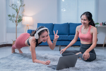 Two sporty asian women are enjoying a workout together at home, following an online fitness class on a laptop and smiling