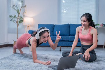 Two sporty female friends wearing headphones and sportswear are enjoying online fitness training together at home, doing plank exercises and following instructions from a laptop