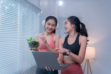 Two young sporty women wearing headphones and sportswear are eating a healthy salad and using a laptop together at home, enjoying a healthy lifestyle and online resources