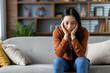 © Liubomir - Asian woman sitting on sofa in cozy home, looking thoughtful. Casual attire suggests relaxed setting. Neutral expression hints at contemplation, reflection in comfortable environment.