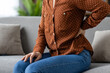 © Liubomir - Woman experiencing back pain sitting on sofa, holding lower back. Wearing brown polka dot shirt and jeans, she shows discomfort, possibly indicating health issues like pain or injury.