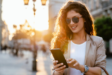 Wall Mural - A stylish Caucasian woman in her mid-adulthood is engrossed in texting on her smartphone, wearing a chic blazer and sunglasses, with the glow of the city illuminating her.