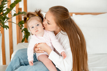 a mother with a baby daughter, a mother gently hugs her baby girl kissing, maternal love and care, a happy family with a baby on the bed at home in the bedroom.