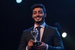 © Maelgoa - A young Middle Eastern businessman in a fitted suit, proudly accepting an award on stage, spotlight on him, with a confident smile, medium close-up showing his hands and trophy 3