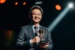 © Maelgoa - A middle-aged Asian businessman in a formal suit, standing on stage, holding an award, smiling proudly under the spotlight, medium close-up showing his hands and trophy 1