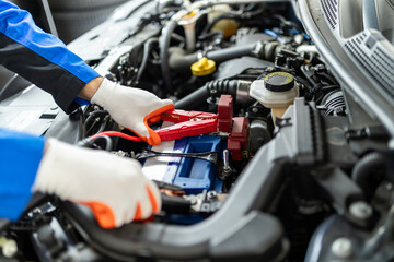 Mechanic jump-starting a vehicle battery in a workshop during daytime with tools and safety gear