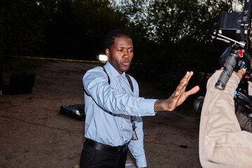 Dramatic portrait of African American man as police officer stopping news reporters and cameras at crime scene, shot with flash