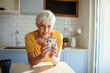 © Marko Geber - Senior woman using smartphone while drinking morning coffee in kitchen