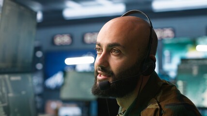 Poster - Middle Eastern military dispatcher relays info from army intelligence analysts to troops on front lines, close up. Radio communications operator discussing with colleague using headset mic, camera A