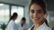 © sirisakboakaew - Beautiful young doctor in a healthcare environment, smiling warmly, with a team member working in the background, creating a professional yet friendly ambiance