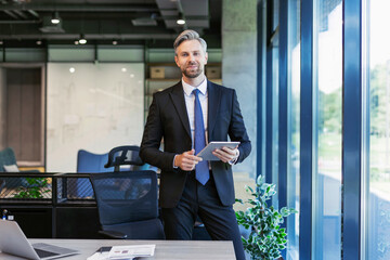 Portrait of smiling middle aged businessman standing with digital tablet at corporate office.