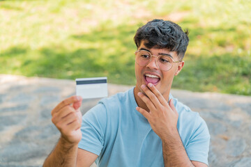 Wall Mural - Young hispanic man holding a credit card at outdoors with surprise and shocked facial expression