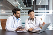 © Liubomir - Doctors in professional attire, stethoscopes visible, collaborate using laptop and tablet in medical office. Discussing patient care with focused expressions, demonstrating teamwork technology use.