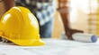 © DuangphonKPR - A yellow hard hat rests on construction plans, with a person in a plaid shirt focused on the project, symbolizing the construction industry.
