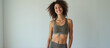 © Roman - A young woman with curly brown hair is smiling at the camera. She is wearing a green sports bra and green leggings. She is standing against a white wall. She has a toned physique and is looking confid