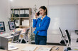 © Parichat - Young businesswoman standing in modern office using smartphone, smiling while talking on mobile phone, computers with data analysis charts on screens in background