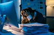 © Parichat - Exhausted businesswoman suffering from headache or migraine, holding head in hands while sitting at desk with computer and stack of paperwork, working late at night in office
