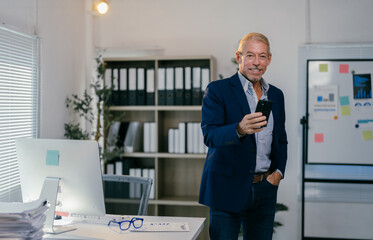 Smiling senior businessman using smartphone in modern office with hand in pocket, standing near desk with computer and documents, shelves with folders and whiteboard with graphs