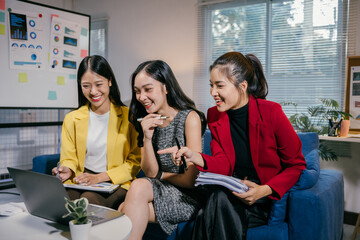 Poster - Three cheerful asian businesswomen are sitting on a sofa in a modern office, collaborating on a project using a laptop and documents, sharing a moment of laughter and positive teamwork
