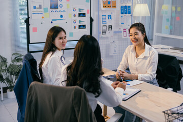 Poster - Three asian businesswomen are discussing marketing strategy during a meeting in a modern office, analyzing charts and reports on a wooden table