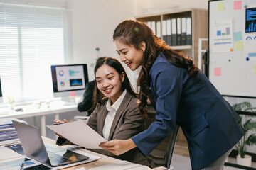 Aufkleber - Two asian businesswomen are smiling while working together in a modern office, using a laptop and reviewing documents, demonstrating teamwork and collaboration