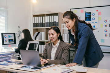 Poster - Two asian businesswomen are working together, using a laptop in a modern office to discuss a project, with charts and graphs visible on the screen and printed documents on the desk