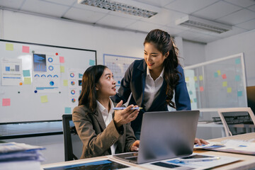 Poster - Two asian businesswomen are discussing over financial charts using laptop in a modern office with whiteboard full of sticky notes in the background