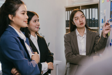 Poster - Asian businesswomen wearing suits are discussing strategy during an office presentation meeting, analyzing charts and graphs on a whiteboard