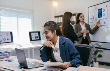 Aufkleber - Young asian businesswoman smiling and working on laptop while her colleagues discussing charts on whiteboard during a meeting in modern office