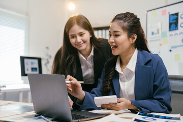 Aufkleber - Two asian businesswomen discussing financial data with a laptop and calculator in a modern office, surrounded by charts and graphs