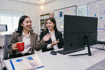 Poster - Two asian businesswomen are discussing a project in the office, looking at computer screen and smiling, one of them is holding a red mug