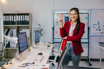 Poster - Asian businesswoman stands confidently in her modern office, holding a sticky note and smiling, surrounded by computers displaying charts and graphs