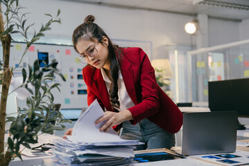 Poster - Young businesswoman wearing eyeglasses and red jacket searching through stack of paperwork at office desk with laptop and charts