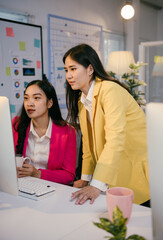 Aufkleber - Two young businesswomen wearing colorful blazers are collaborating on a project, focusing on a desktop computer in a contemporary office setting with charts and notes visible in the background