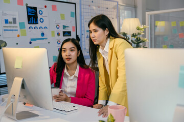 Aufkleber - Two young businesswomen are collaborating on a project, analyzing data on a desktop computer in a bright, modern office with charts and graphs displayed on a whiteboard