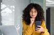 © Liubomir - Businesswoman with curly hair holding smartphone in office setting. Reflective expression conveys thoughtfulness and focus. Ideal for themes of technology, professionalism, and contemporary work.