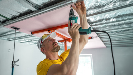 Wall Mural - The worker screwing plasterboard to the ceiling. He is using special electric screwdriver and drywall lift. A red plasterboard improve the fire resistance of ceiling structures.