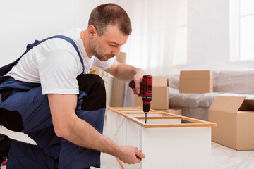 Fototapete - Furniture Assembly Service. Man In Blue Coverall Installing And Assembling Wooden Shelf Using Electric Drill Indoors. Side View Shot Of Carpenter Fixing Furniture At Work. Carpentry Concept