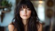 © peppastocker - A woman with long dark hair and bangs poses indoors with a serene expression, accentuated by soft natural lighting creating a calm and reflective ambiance.