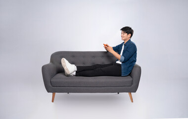 Wall Mural - Full body image of young Asian man using phone sitting on sofa on white background