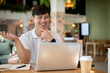 © bongkarn - An Asian man is working remotely from a coffee shop, sitting at an indoor table with his laptop.