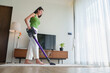 © eggeeggjiew - Asian Woman vacuuming the living room floor with a cordless vacuum cleaner for housekeeping housework and chores, focusing on keeping her home clean and organized.