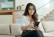 © Rapid Stock - Happy and smiling young Latina woman sitting on a sofa holding her smartphone in a brightly lit beautiful livingroom with a bokeh background