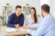 © Studio Romantic - Cheerful family couple preparing to sign contract at realtor office. Professional real estate agent sitting at desk with happy clients, finalizing details of deal and showing where to put signature.