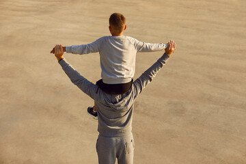 Back view of child boy in sportswear sitting on fathers shoulders holding hands and looking into the distance. Father with son enjoying time together after sport workout outdoors. Family sport.