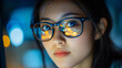 © 1by1step - A close-up of an Asian woman's face, wearing glasses and looking at the camera with bright lights reflecting on her eyeglasses in front of a computer screen