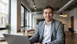 © Nattawat - A confident businessman in a suit, smiling, works on a laptop in his office